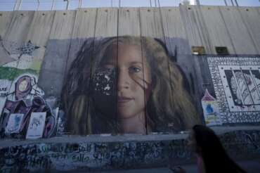 A tourist pauses in front of a mural depicting Palestinian activist Ahed Tamimi on Israel’s controversial separation barrier in the West Bank city of Bethlehem, Sunday, June 19, 2022. The summer of 2022 can feel as if the coronavirus pandemic is really over — mask rules and testing requirements are lifting in many countries, including the United States. Full recoveries are generally not expected until at least 2024. (AP Photo/Maya Alleruzzo)