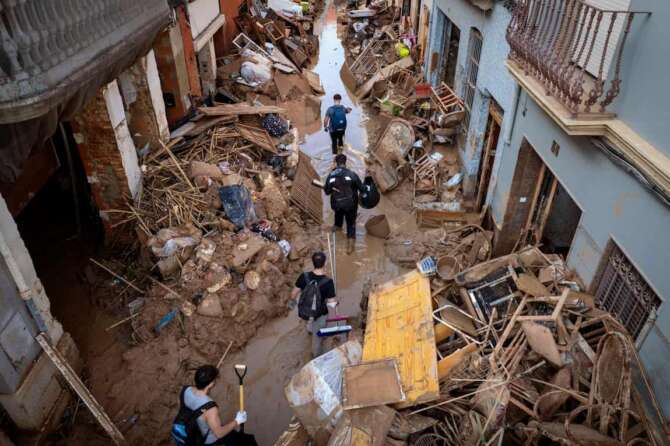 People walk through a street with piled furniture and rubbish on the sides in an area, affected by floods, in Paiporta, Valencia, Spain, Tuesday, Nov. 5, 2024. (AP Photo/Emilio Morenatti)