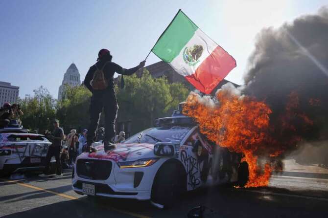 A protester stands on a burning Waymo taxi near the metropolitan detention center of downtown Los Angeles, Sunday, June 8, 2025, following last night’s immigration raid protest. (AP Photo/Eric Thayer) Associated Press/LaPresse